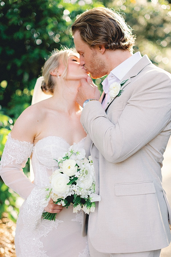Wedding kiss portrait of bride and groom kissing as she holds a white rose bouquet, framed by veil and sunlit trees bokeh