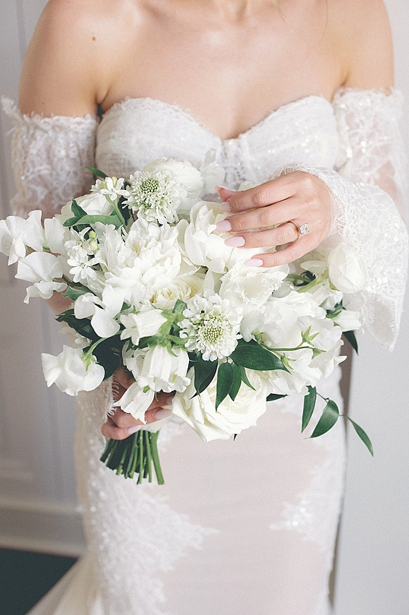 Bridal bouquet of white wedding bouquet blooms with greenery held by a bride in an off-the-shoulder lace dress, ring visible in soft window light