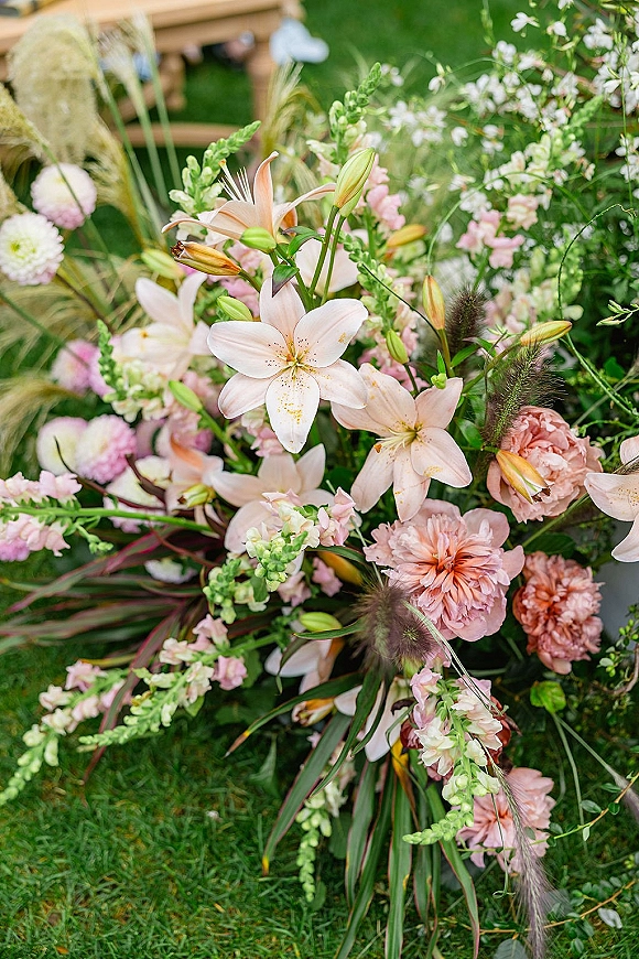 Wedding floral arrangement of ceremony aisle flowers with pink lilies and blush blooms set on a grass lawn beside a wooden bench