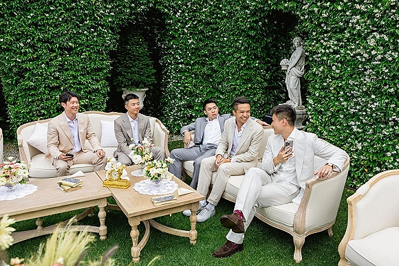 Groomsmen portrait of men in light suits lounging on an outdoor sofa, with boutonnieres and a floral centerpiece by a hedge wall