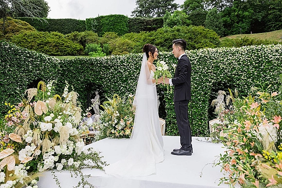 Wedding ceremony moment as couple exchanging vows on a garden platform, bride in strapless dress and long veil holding bouquet, groom in tuxedo amid white florals