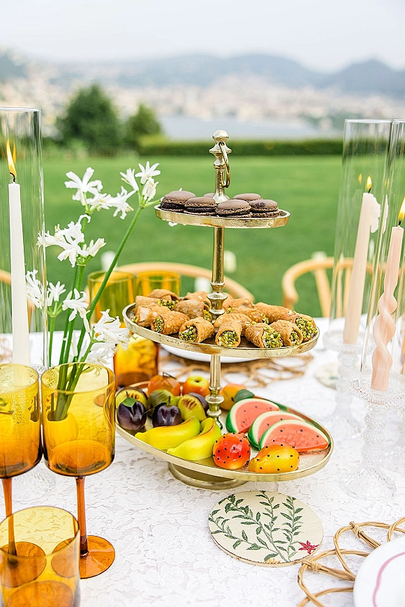 Wedding dessert table with tiered dessert stand of macarons, pastries, and fruit on lace tablecloth, with taper candles on a lawn with hills and water view