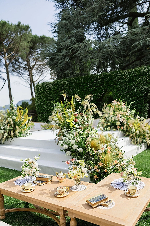 Outdoor ceremony setup with garden wedding ceremony florals around a white platform stage and lounge seating, plus vintage teacups on a coffee table