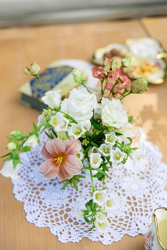Wedding bouquet with white flowers and blush bloom beside a vintage perfume bottle and book on a wood tabletop, with lace doily accent