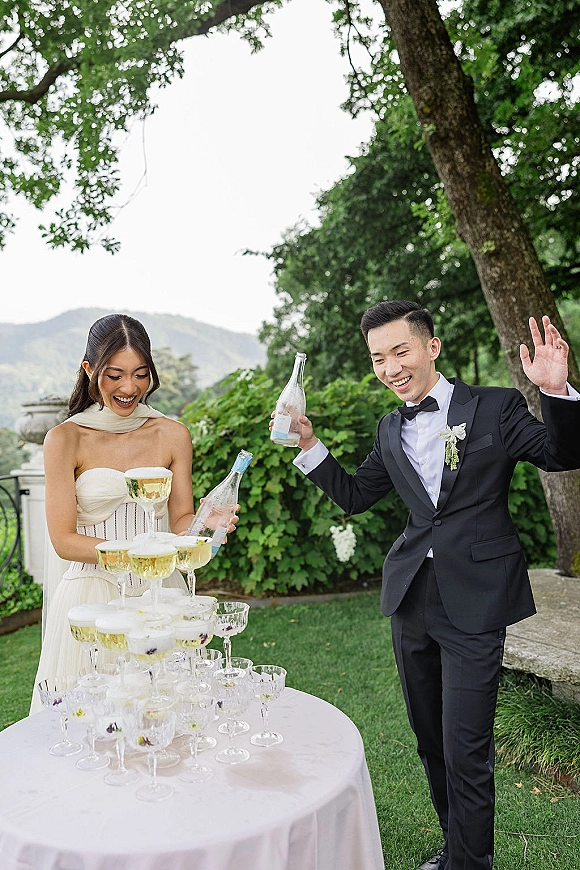Champagne tower moment as bride in strapless dress and groom in tux pour from bottles into coupe glasses on a white table outdoors