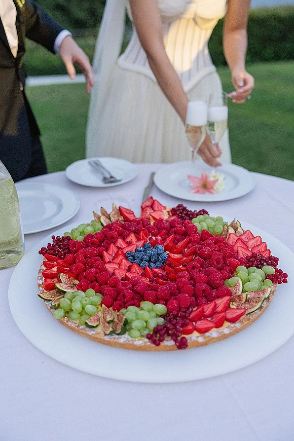 Wedding dessert table with fruit tart and mixed berries, champagne flutes and bottle on a white tablecloth, set on an outdoor lawn