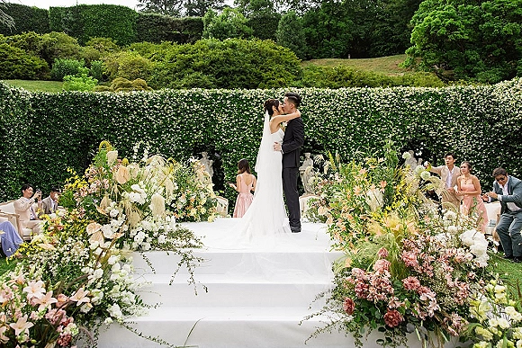 Wedding kiss as bride in a simple wedding dress and veil kisses groom in a black suit on a garden ceremony platform with guests