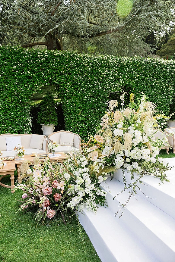 Ceremony floral decor with wedding ceremony flowers framing a white platform, pampas grass and greenery beside a cream lounge on a hedge-lined lawn