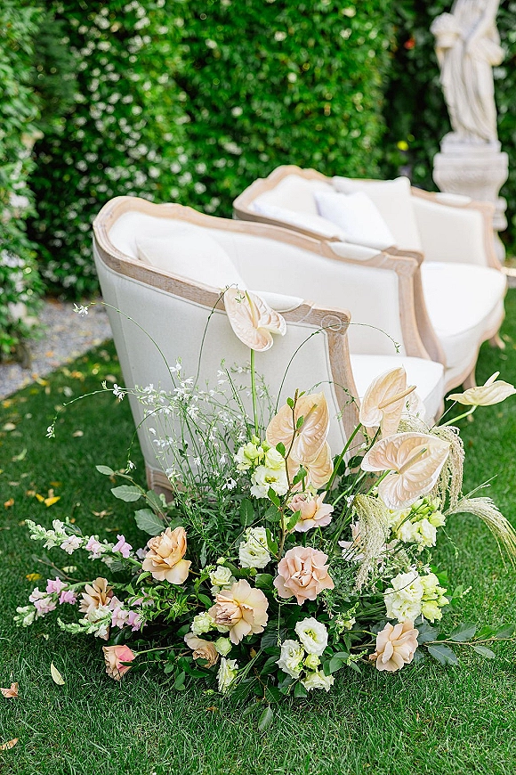 Wedding ceremony seating with an ivory upholstered loveseat, white cushions, and blush florals with greenery on a garden lawn by a hedge