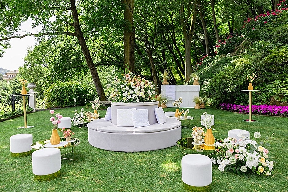 Wedding lounge setup with a round sofa and mirrored coffee tables, styled with floral arrangements on a garden lawn by a stone balustrade