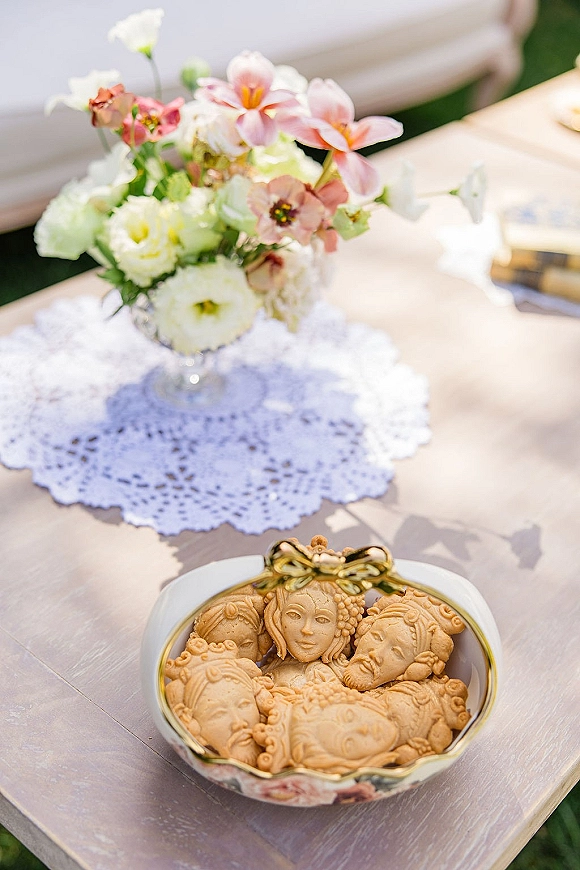 Wedding favors with custom wedding cookies on a ceramic dish, ribbon detail, and small floral arrangement on lace doily atop pink linen outdoors