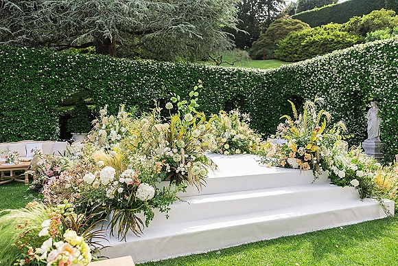 Wedding ceremony setup with outdoor ceremony platform, featuring a white stage with meadow florals, pampas grass, and a lounge sofa by a hedge wall.