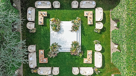 Outdoor ceremony setup with a garden ceremony platform, white stage and floral corners, plus lounge sofas and table settings on a grassy lawn
