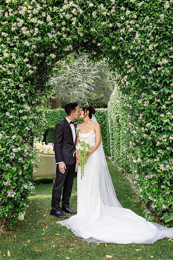 Wedding kiss portrait of bride and groom kissing under a flowering vine archway, her veil trailing as he stands in a black tuxedo