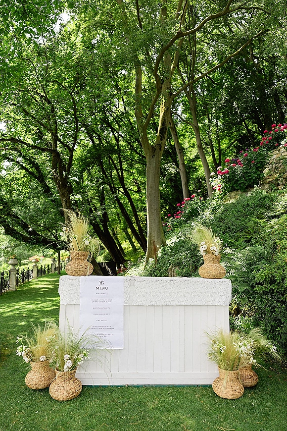 Wedding bar setup with a white bar and menu sign on a lace tablecloth, styled with pampas grass and flowers on a garden lawn