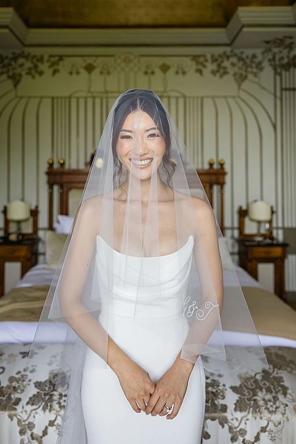 Bridal portrait of a bride wearing veil over her face, smiling in a strapless wedding dress beside a bed in a softly lit suite
