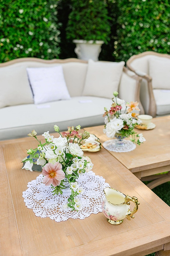 Reception lounge decor with floral arrangements on a wooden coffee table, vintage teacups and lace doilies beside a sofa in a garden hedge setting