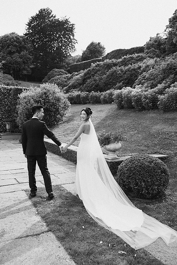 Couple portrait of bride and groom holding hands, bride glancing back with wedding veil train flowing on a garden stone walkway