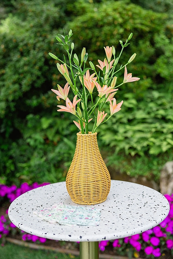 Wedding centerpiece in a wicker vase with peach lilies on a round terrazzo cocktail table, set against garden greenery and stone edging