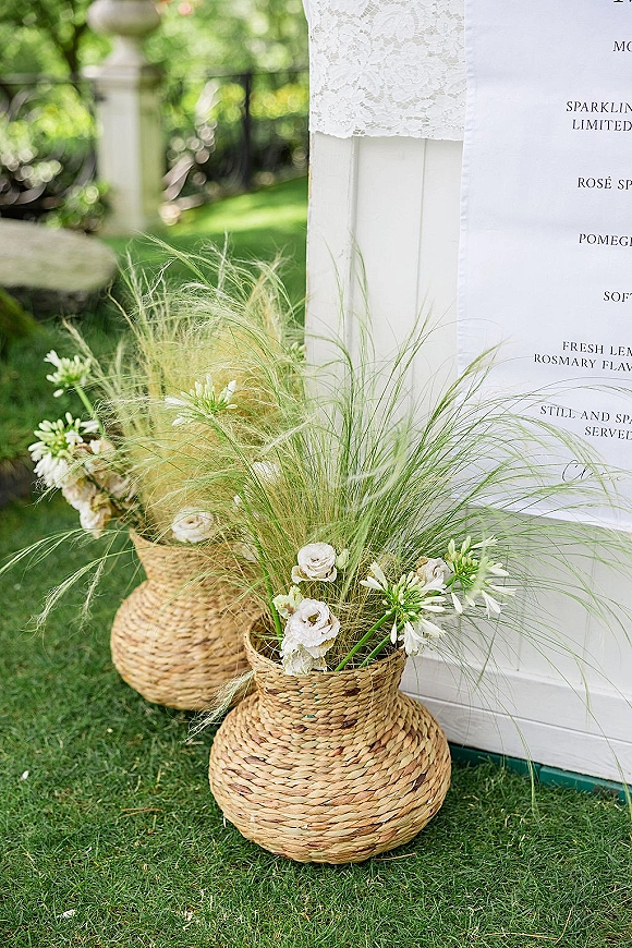 Wedding signage with welcome sign florals on a white board, lace-draped baskets and pampas grass on a garden lawn by a metal fence