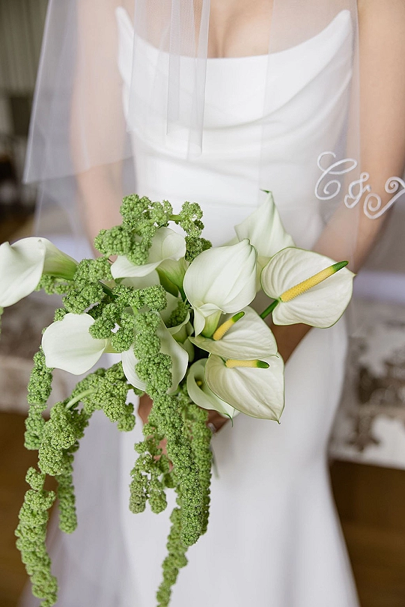 Bridal bouquet of white calla lilies and anthurium with cascading greenery, held by bride in strapless gown and veil in a neutral room