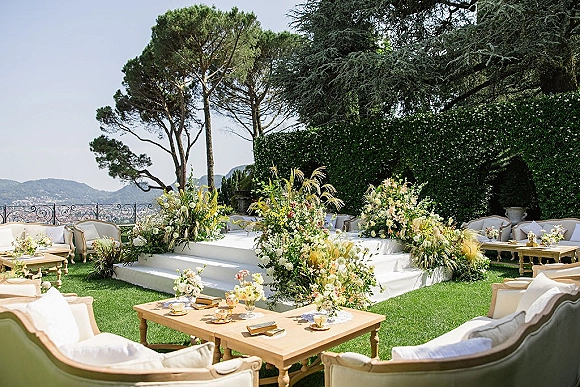 Outdoor ceremony setup with a white platform stage, asymmetrical pastel florals and lounge seating on a manicured lawn by tall trees and hills