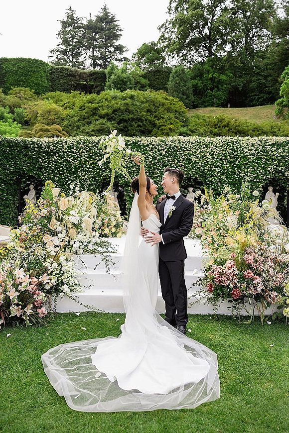 Couple portrait of bride and groom embrace as she lifts a white bouquet, veil trailing on garden ceremony steps with hedges and statues