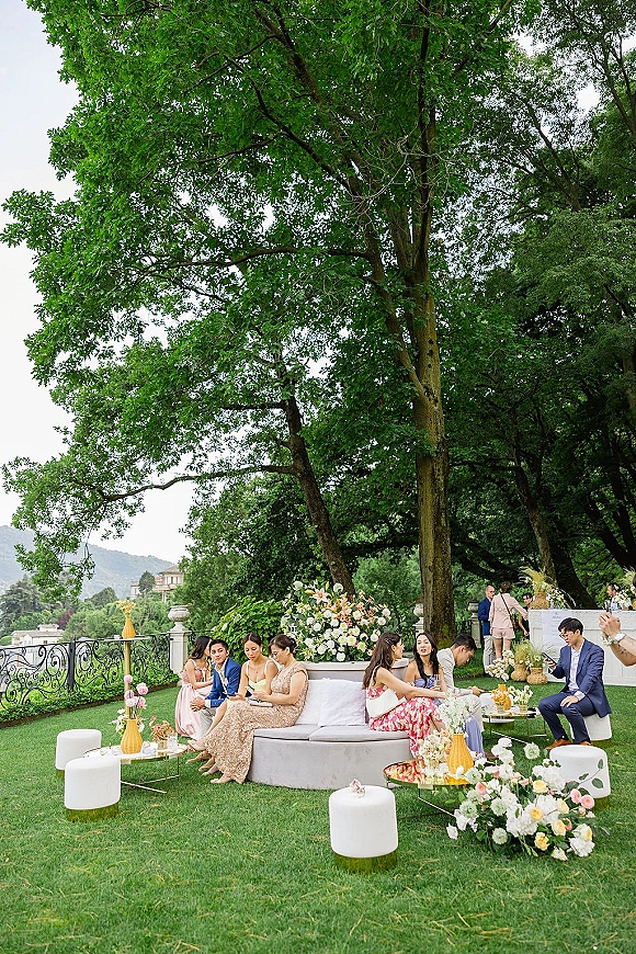 Outdoor wedding lounge with white sofa and ottomans around cocktail tables, accented with candles and florals on a tree-lined lawn