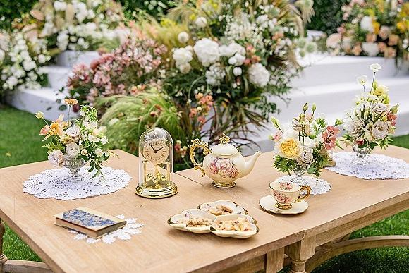 Wedding tea table set with a vintage teapot, teacups and cookies on a wood table, with lace doilies and a floral ceremony backdrop on the lawn