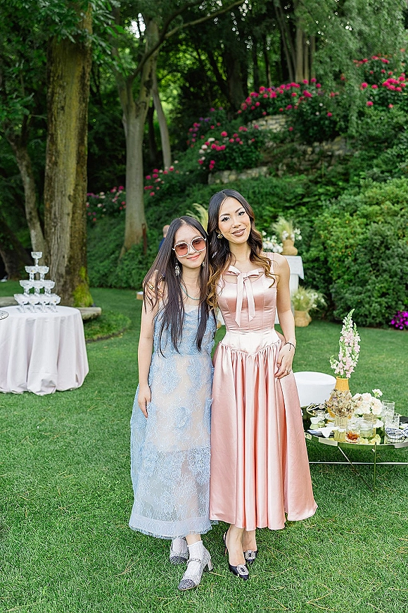 Wedding guest portrait of two women in a pink satin dress and blue lace dress on a garden lawn near a champagne tower display