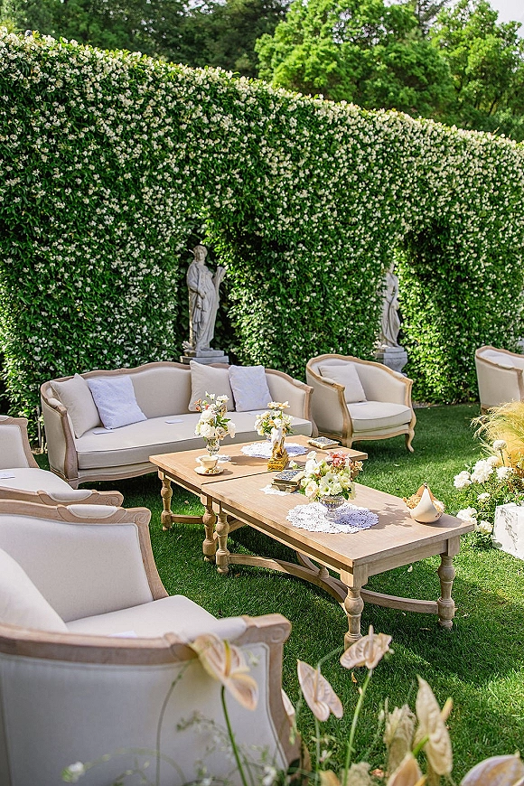 Outdoor lounge setup with wedding lounge seating, vintage sofa and armchairs around a wooden coffee table with floral centerpieces by a flowering hedge wall