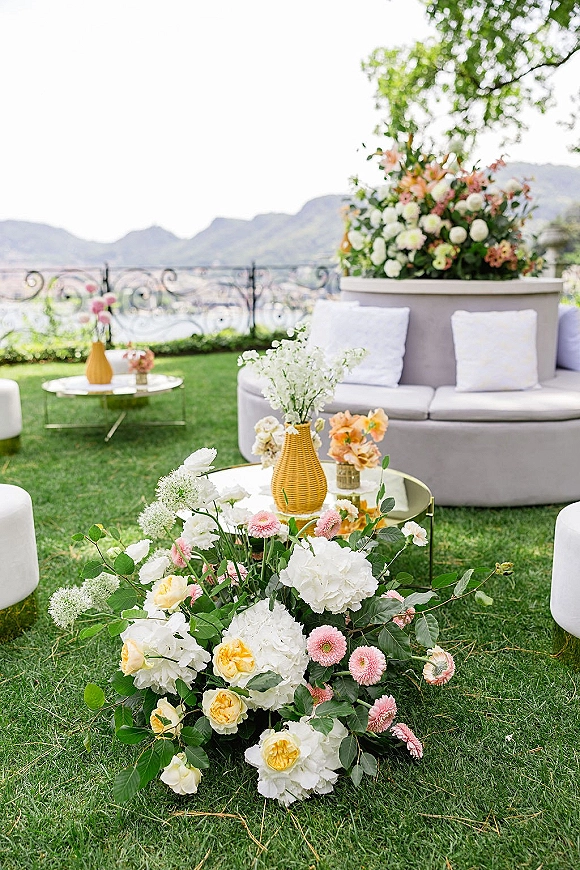 Outdoor lounge seating with a round sofa, throw pillows, and glass coffee tables with yellow vases, set on a lawn by a lake and mountains