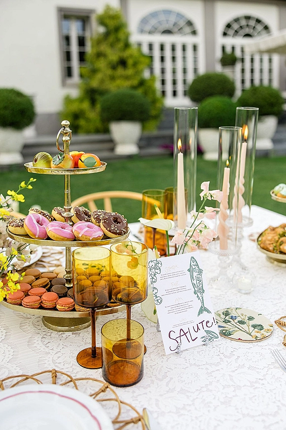 Wedding dessert table with donut dessert display on tiered stands, macarons, fruit, taper candles and amber goblets on a lace cloth in a garden patio setting