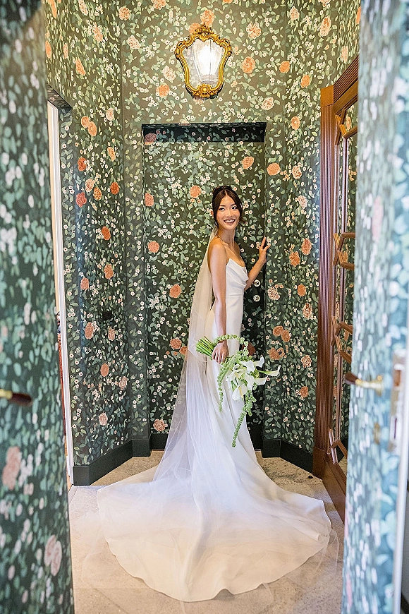 Bridal portrait of bride holding bouquet in a strapless wedding dress with long veil, standing in a doorway against floral wallpaper hall