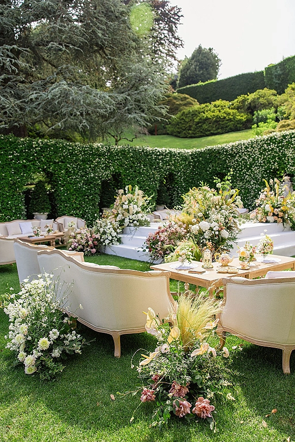 Wedding lounge setup with cream sofas, wooden coffee tables, and tea service decor, framed by floral meadow and greenery hedge backdrop on a lawn