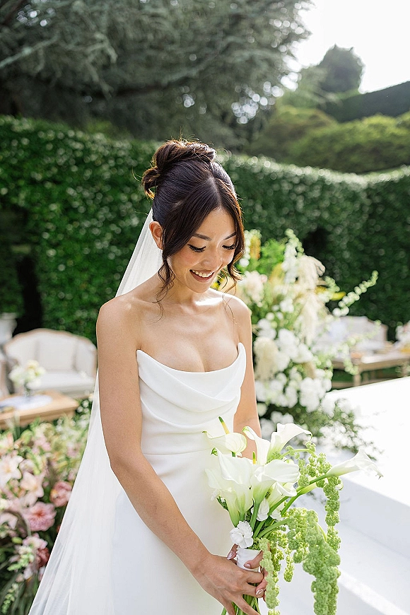 Bridal portrait of a smiling bride holding bouquet with a white calla lily bridal bouquet, wearing a veil in sunlit garden hedges