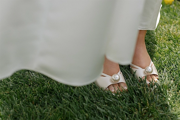Bridal shoes with pearl wedding heels detail, open-toe white sandals beside a white dress hem resting on a grass lawn