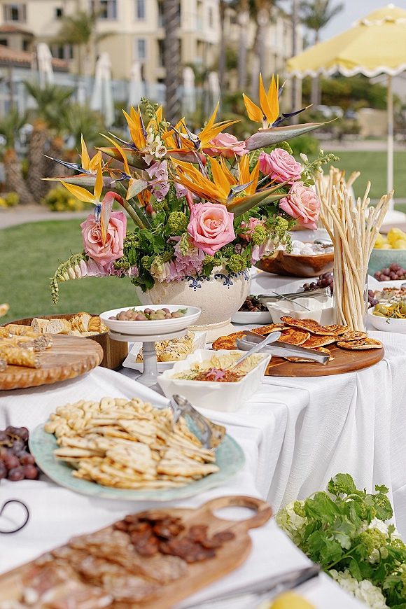 Wedding appetizer table set for cocktail hour with wood boards of flatbreads, dips, olives and grapes, plus bird of paradise centerpiece on a lawn patio