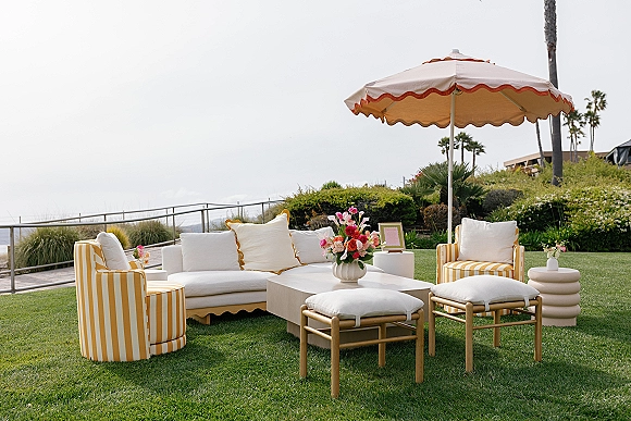 Outdoor lounge seating with a patio umbrella, white sofa, striped armchairs and floral centerpiece on a coastal lawn under overcast skies