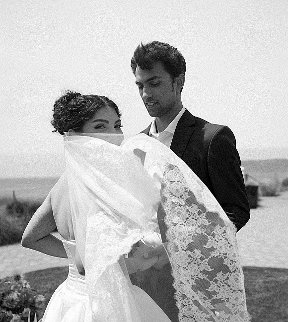 Couple portrait in black and white, groom in dark suit holding bride with lace veil over her face by an ocean coastline backdrop