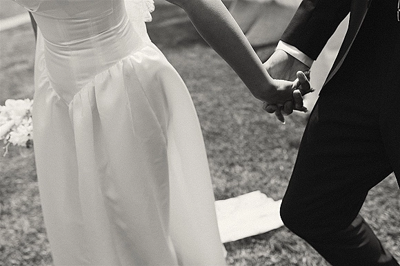 Ceremony moment with wedding hand holding, bride and groom hands close up showing ring, suit cuff, and dress train on grass lawn
