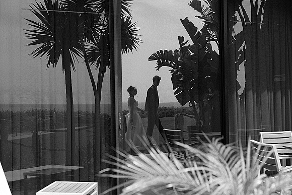Couple portrait in a black and white wedding photo, bride in strapless dress holding bouquet beside groom, reflected on glass terrace with palms and ocean horizon