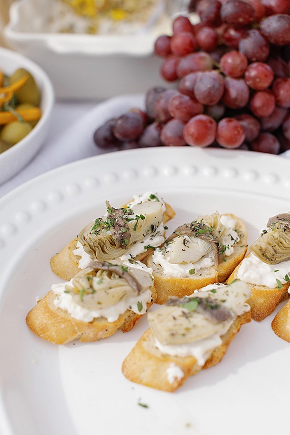 Wedding cocktail appetizers arranged on a white platter with ricotta and artichoke crostini, herbs, grapes, and olives on a catering table