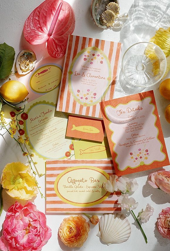 Invitation suite wedding invitation flat lay with striped envelope, RSVP and menu cards, gold rings, pearls, lemons, and flowers on white in sunlight