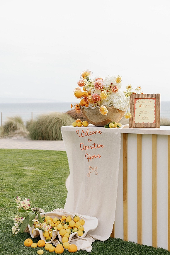 Cocktail hour decor with a wedding aperitivo bar, framed drink menu, citrus and floral bowl vase centerpiece on a runner by the ocean lawn