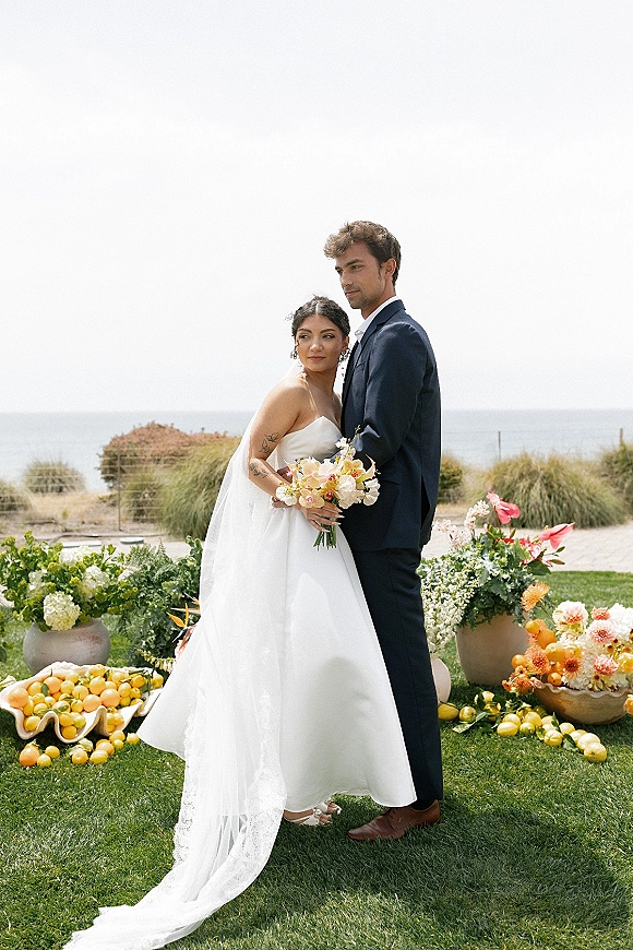 Couple portrait of an outdoor bride and groom embracing, bride in strapless dress holding bouquet on an oceanfront lawn with coastal grasses
