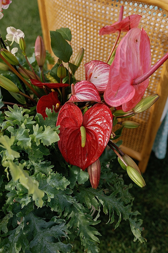 Wedding floral arrangement with anthurium wedding flowers, lily buds and lush greenery draped over a rattan chair on a grass lawn
