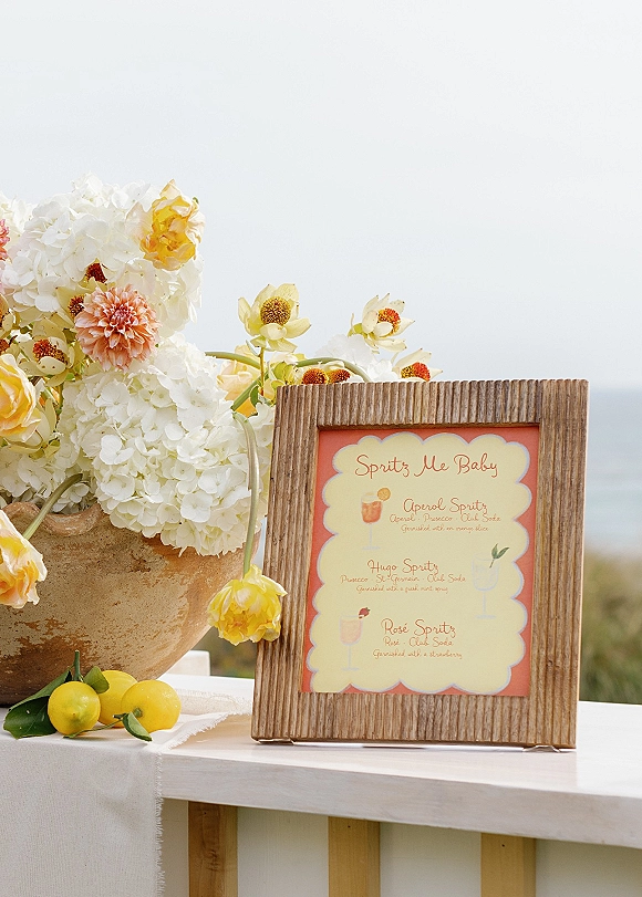 Wedding bar menu framed sign displaying a signature cocktail menu beside hydrangeas, peach dahlia, and lemons on an outdoor table