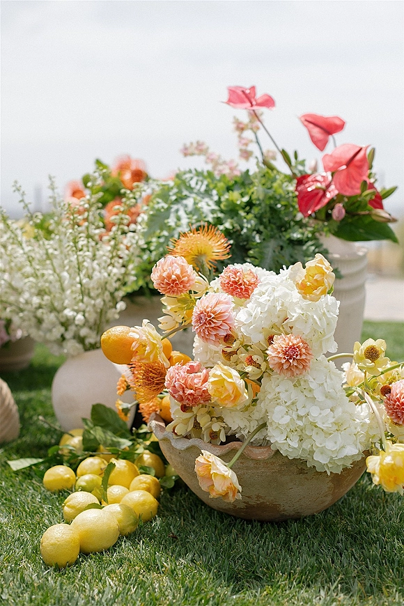 Wedding florals with citrus wedding flowers in a stone bowl, featuring hydrangeas, dahlias, roses, greenery, lemons and limes on a lawn under open sky