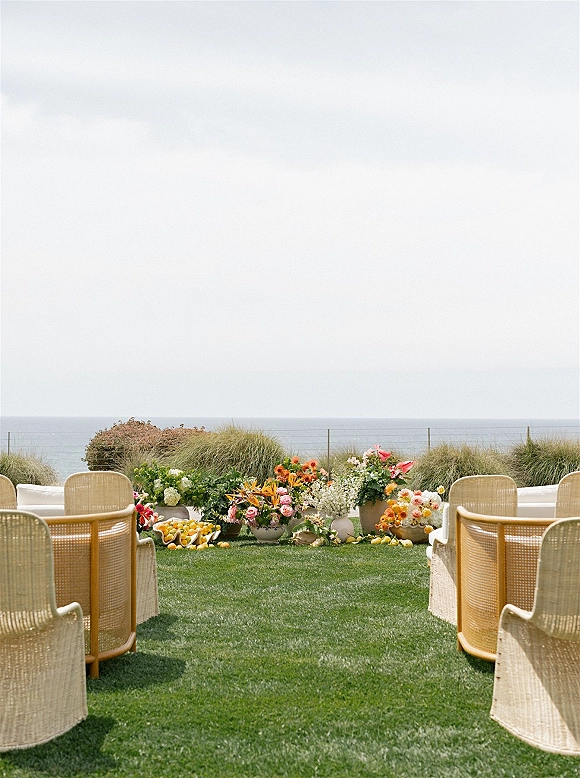 Ceremony aisle decor with wicker chairs and ground florals in ceramic planters accented by citrus, set on a lawn with ocean view